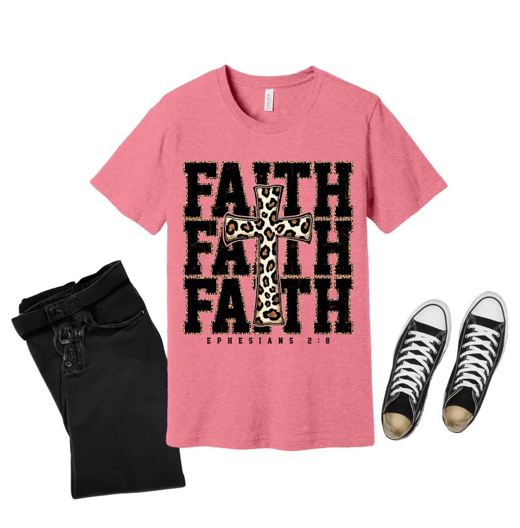 Pink t-shirt with 'FAITH' and cross design on a wooden surface with black pants and white sneakers.